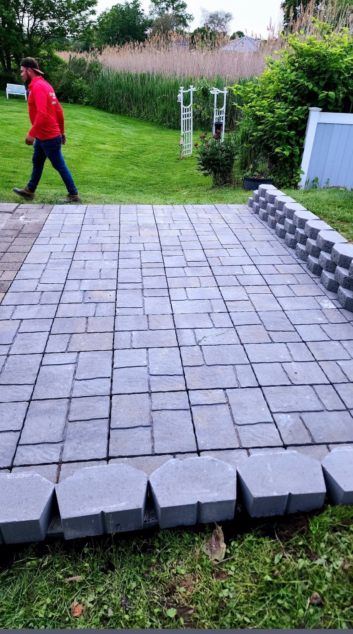 Gray paver patio in garden with person in red jacket walking on grass in background