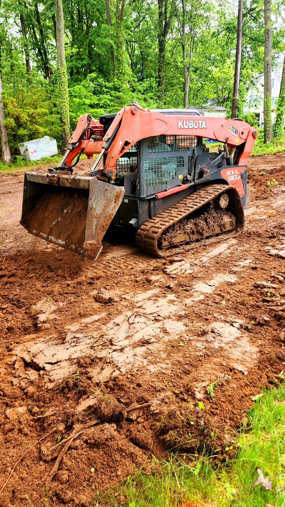 Orange Kubota skid-steer loader with front bucket on a freshly graded dirt work site surrounded by green trees during a residential project